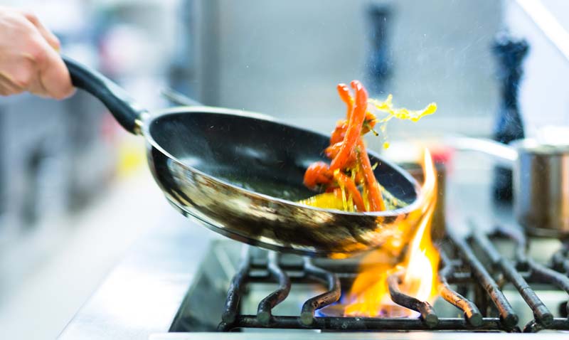 portfolio image: professional stock photo of a pan, inside it peppers being flipped, over a stove with flickering flame.