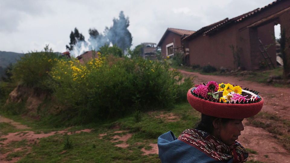 Fine art photo by Prantik Mazumder, Peruvian woman with flowers collected in her hat in foreground, humble architectural form and foliage in middle ground, some smoke and tress in background.