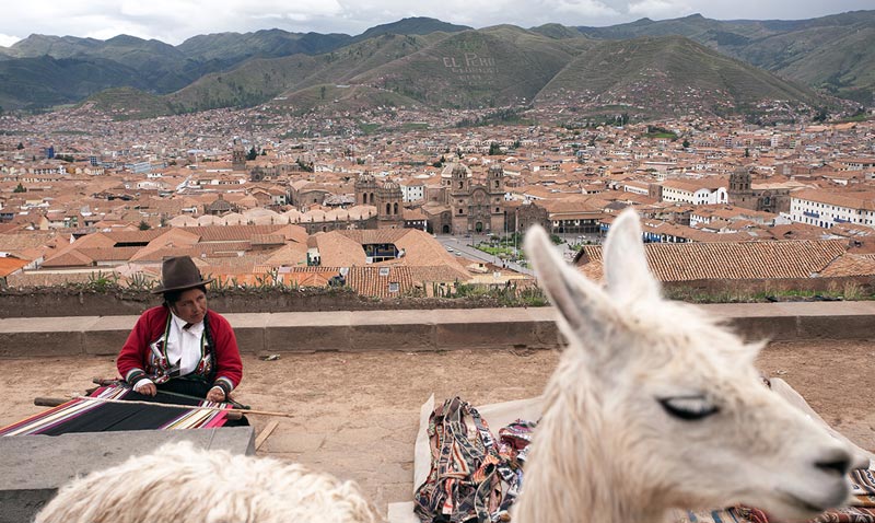 Fine art photo by Prantik Mazumder, Peru mountains in background, woman working on a tapestry in middle ground, a blurred white mule in foreground.